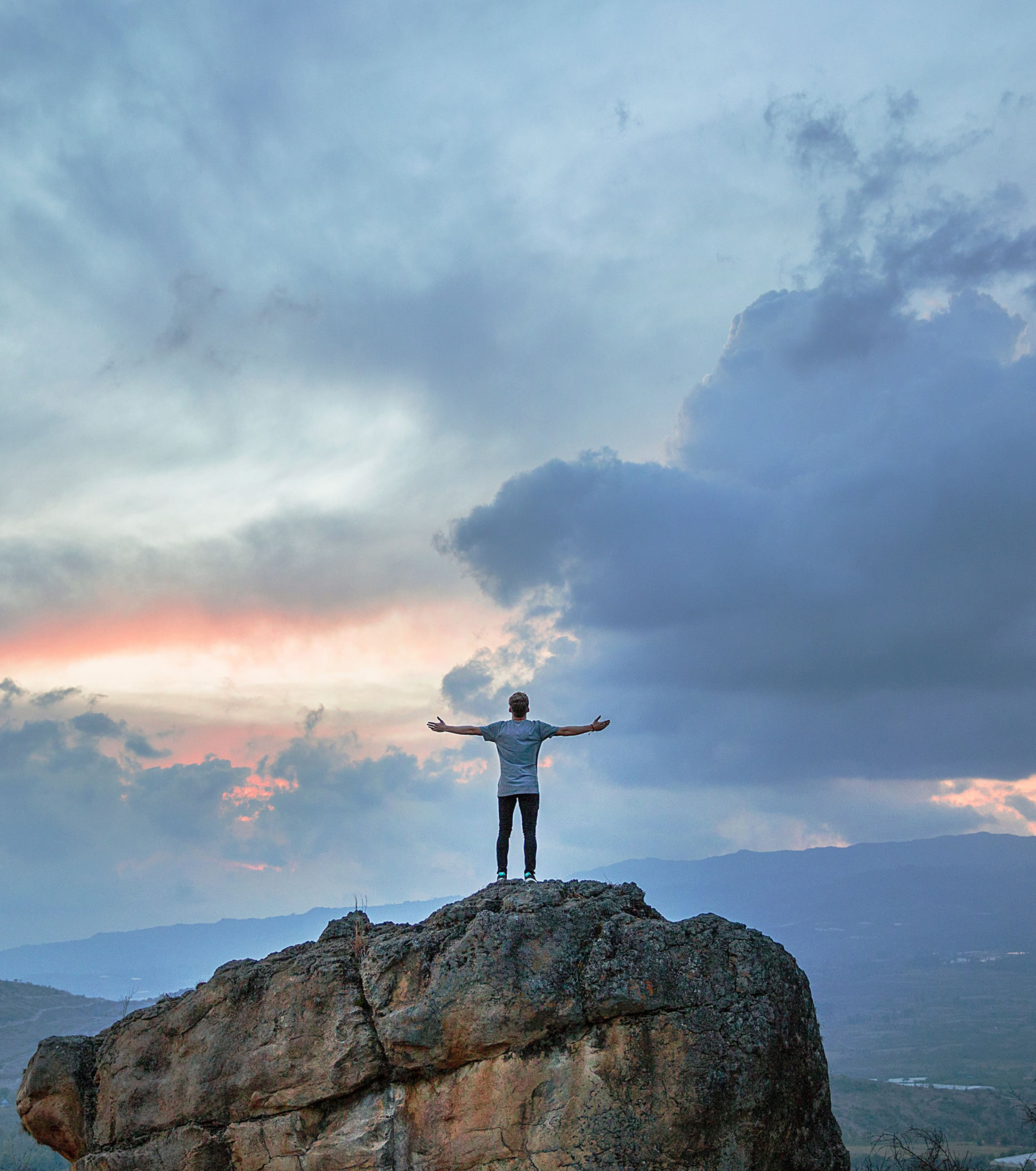 man standing on top of a large rock face with hands outstretched towards a colourful bright evening sky